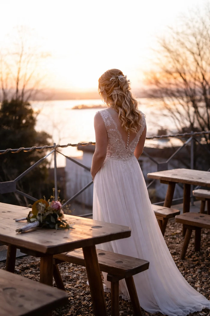 Braut-Editorial bei Sonnenuntergang am Wasser. Blondes Model im fließenden weißen Brautkleid mit elegantem Spitzenrücken, natürliches Abendlicht und romantische Atmosphäre.
