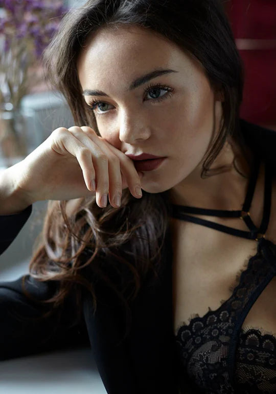 Editorial-Porträt eines Models mit schwarzen Haaren und grünen Augen. Sie trägt elegante schwarze Hunkemöller-Dessous und ein natürliches Abend-Make-up mit strahlendem Glow. Offene Haare, weiches Fensterlicht und eine elegante, zeitlose Ausstrahlung
