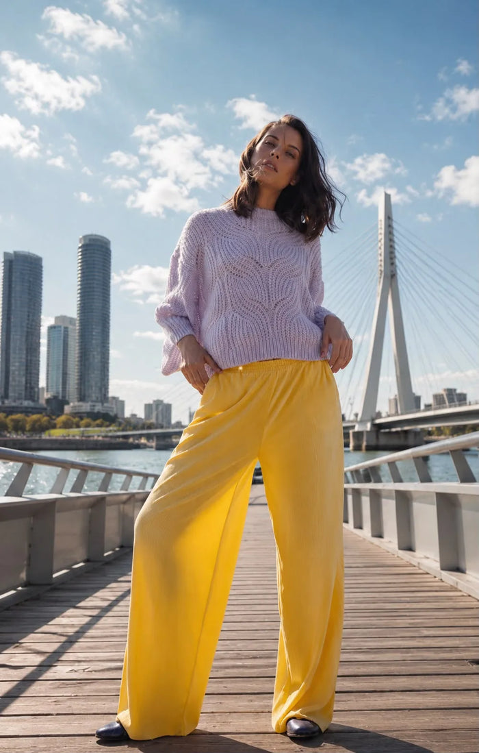 Ganzkörperaufnahme eines Models auf einer Brücke mit Wasser, Skyline und moderner Stadtarchitektur im Hintergrund. Das Model trägt eine weite gelbe Hose und einen fliederfarbenen Strickpullover von Designer Remix Charlotte Eskildsen sowie flache Schuhe. Schulterlange dunkle Haare, natürliches Make-up und Gegenlicht verleihen dem Bild einen modernen, frischen Fashion-Look.