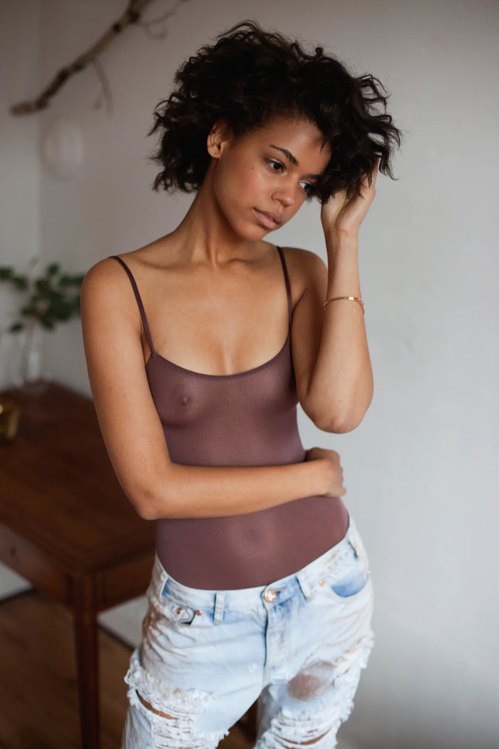 Fashion-Editorial in einem Innenraum mit natürlichem Fensterlicht. Brasilianisches Model mit kurzen schwarzen Locken trägt einen transparenten braunen Spaghetti-Body und hellblaue verwaschene Jeans im Used-Look mit Rissen. Natürliches Make-up, warme Hauttöne und eine ruhige, moderne Atmosphäre verleihen dem Bild einen authentischen Fashion- und Lifestyle-Charakter.