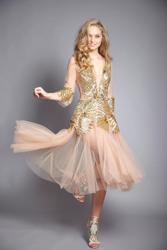 Elegantes gold- und nudefarbenes Abendkleid aus Tüll von Elisabetta Franchi. Blondes Model mit blauen Augen, weichen Locken und natürlichem Make-up trägt passende High Heels. Romantischer, sinnlicher Look mit griechisch inspirierter Eleganz vor grauem Studiohintergrund.