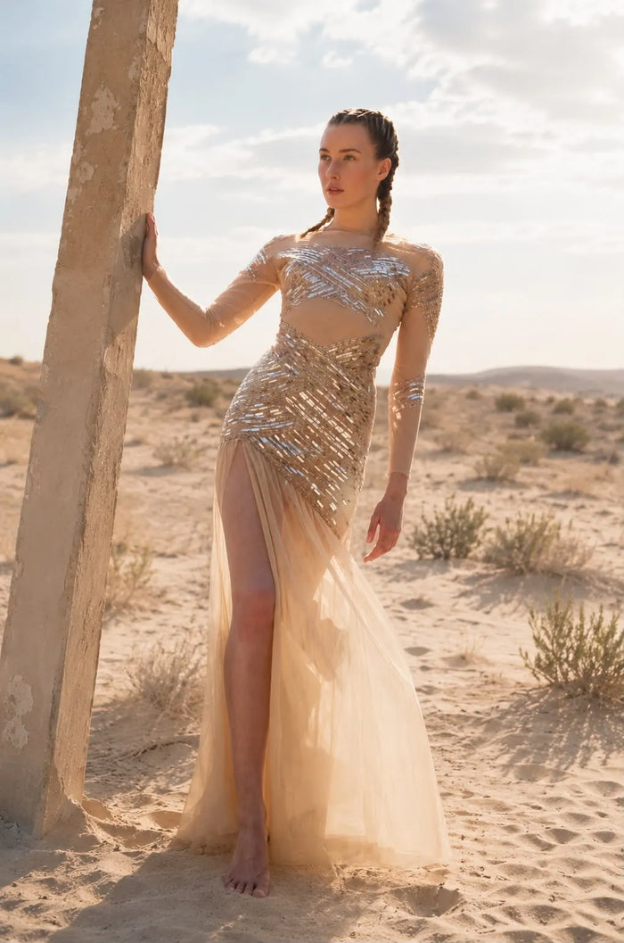 Elegantes gold- und nudefarbenes Abendkleid von Elisabetta Franchi mit transparentem Tüll, goldenen Stickereien und fließender Silhouette. Das Model steht barfuß in einer Wüstenlandschaft im warmen natürlichen Sonnenlicht. Das luxuriöse Kleid erinnert mit seinen goldenen Details an antike, griechisch-römisch inspirierte Eleganz und verleiht dem Bild eine zeitlose, glamouröse Ausstrahlung.