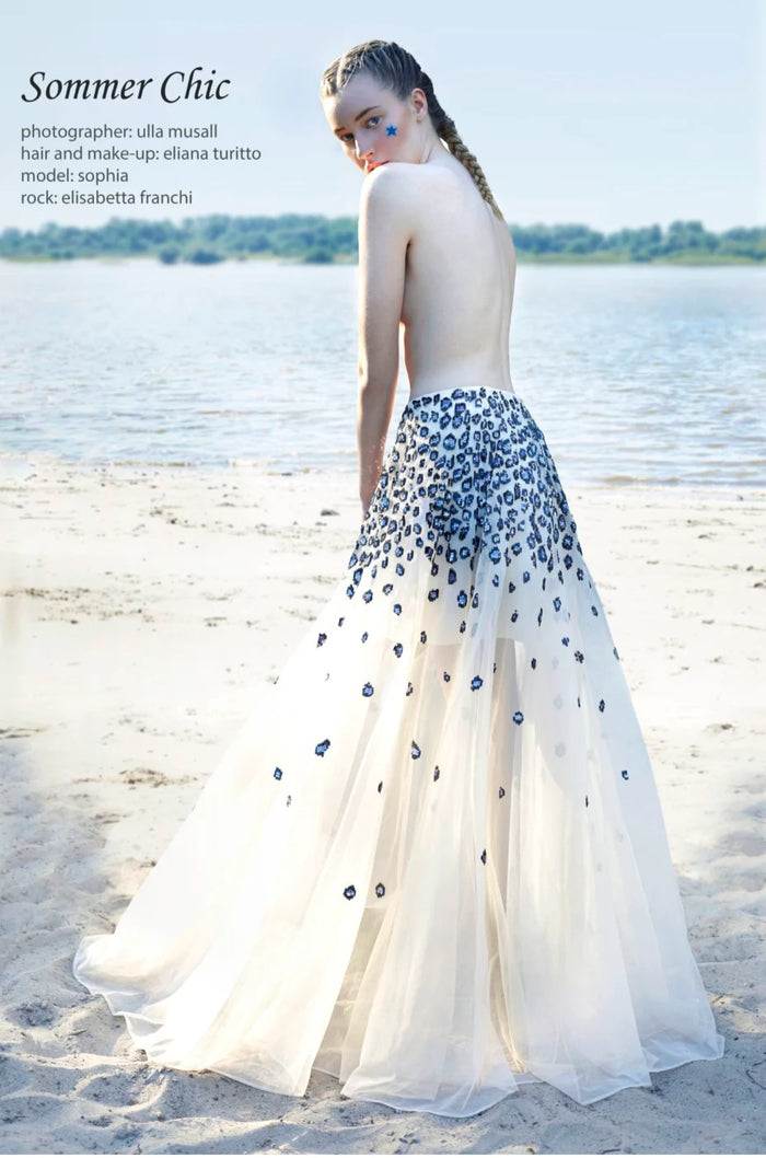 Blondes Model mit geflochtenen Haaren am weißen Sandstrand in einem langen transparenten Tüllrock von Elisabetta Franchi. Der Rock ist mit kleinen dunkelblauen Punkten verziert, die nach unten hin feiner werden. Im Hintergrund sind Wasser und ein grünes Ufer bei natürlichem Tageslicht zu sehen.