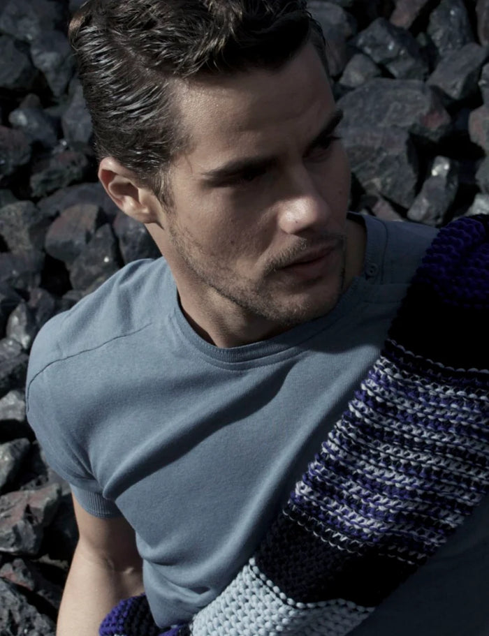 Herren-Fashion-Porträt am Hafen mit natürlichem Sonnenlicht. Model mit kurzen dunklen Haaren trägt ein petrolfarbenes T-Shirt und einen Strickpullover ,über der Schulter. Die Steinmauer im Hintergrund und das seitliche Sonnenlicht verleihen dem Bild einen modernen, maskulinen und zeitlosen Look.
