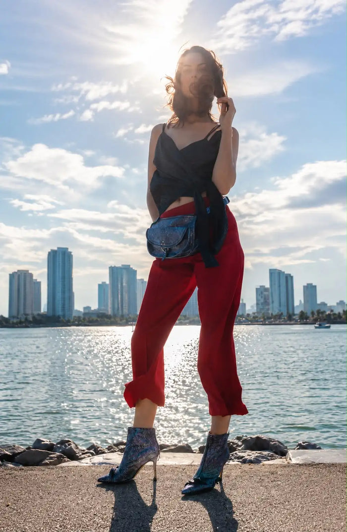 Fashion-Aufnahme an der Miami Waterfront im natürlichen Gegenlicht. Das Model trägt eine rote Hose von Designer Remix Charlotte Eskildsen, ein schwarzes Spaghettiträger-Top, glitzernde Stiefeletten mit Absatz und eine Crossbody-Tasche. Die Skyline im Hintergrund, das Wasser und das warme Sonnenlicht verleihen dem Bild einen modernen, urbanen und eleganten Fashion-Look.