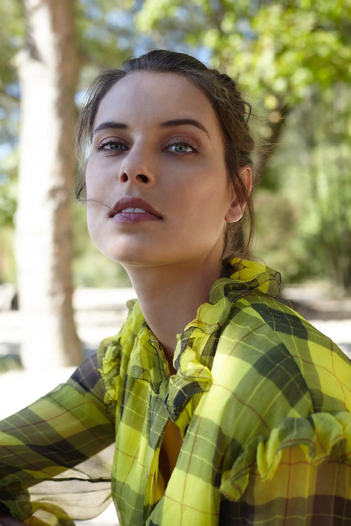 Fashion-Editorial im Freien mit natürlichem Sommerlicht. Das Model trägt ein gelb kariertes Kleid mit Rüschendetails von Designer Remix Charlotte Eskildsen. Fotografiert unter Bäumen mit weichem Tageslicht, natürlichem Make-up und einem eleganten, ausdrucksstarken Blick für einen modernen Fashion-Look.