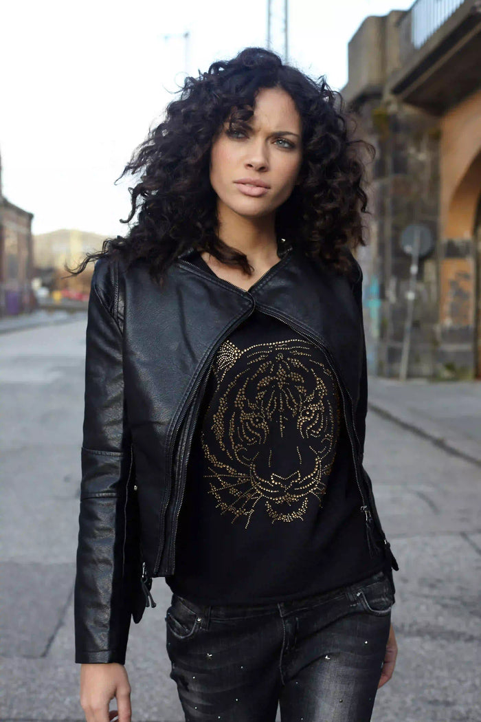 Fashion-Streetstyle mit brasilianischem Model und schulterlangen schwarzen Locken. Das Model trägt eine schwarze Lederjacke über einem Shirt von Miss Goodlife mit goldfarbenem Print sowie eine dunkle Jeans. Natürlicher Look, warme Hauttöne und eine urbane Straßenkulisse verleihen dem Bild einen modernen und selbstbewussten Stil.