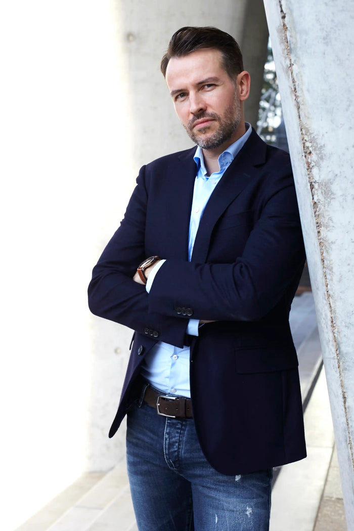Business-Porträt eines Unternehmers im Business-Casual-Stil. Dunkelblaues Sakko, hellblaues Hemd und Jeans vor moderner Architektur. Stilvolles Männerporträt mit natürlichem Licht – ideal für Personal Branding, Business-Websites und LinkedIn-Profil.