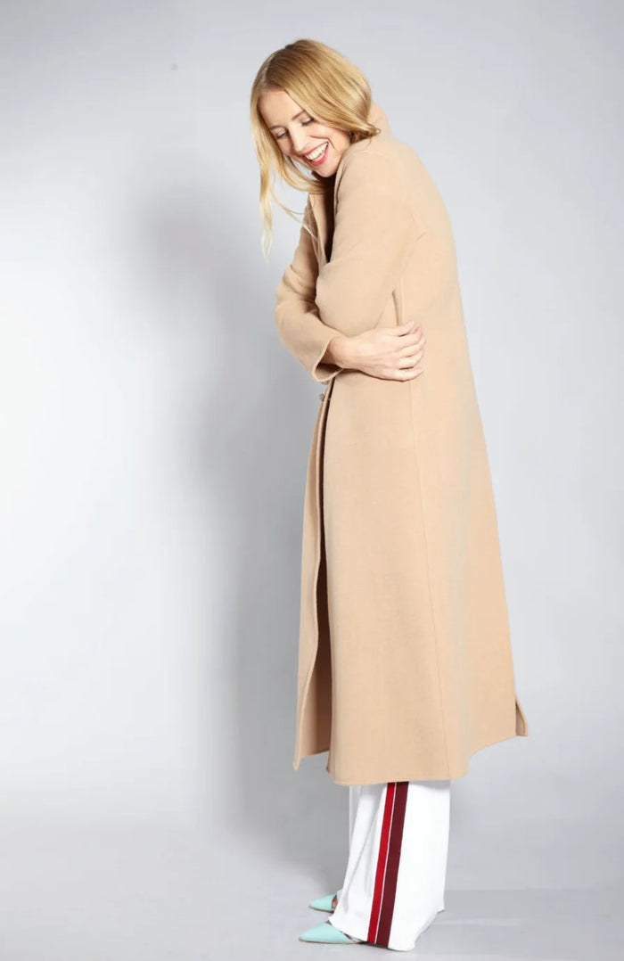 Blondes Model im langen camelfarbenen Mantel mit weißer Hose und hellen Schuhen. Eleganter Winter- und Streetstyle-Look im Fotostudio vor grauem Hintergrund, natürliche Ausstrahlung und sanftes Lächeln.