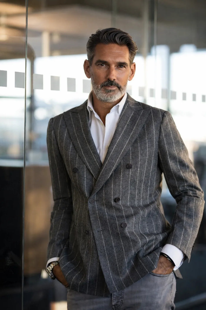 Lifestyle-Business-Porträt eines eleganten Mannes um die 50 mit graumeliertem Haar und gepflegtem Bart. Er trägt einen grauen doppelreihigen Nadelstreifenanzug mit weißem Hemd und posiert selbstbewusst in einer modernen Büro- und Glasarchitektur. Stilvolles Business-Fotoshooting mit natürlichem Licht und zeitloser Eleganz.